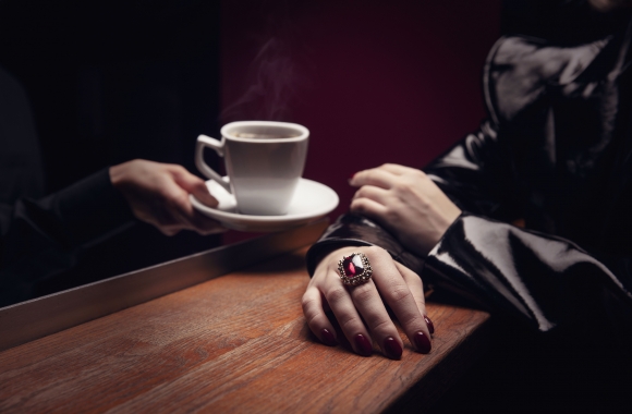 hämärässä valaistuksessa käsi ojentaa valkoisen kahvikupin asiakkaalle jolla on punainen sormus sormessa